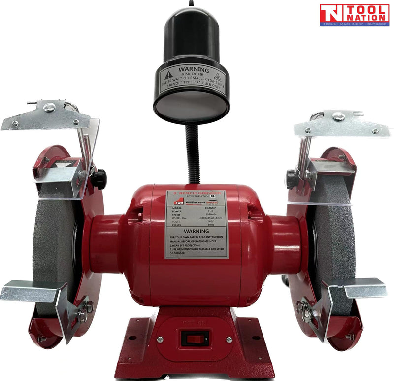 Red bench grinder with two grinding wheels, clear eye shields, metal tool rests, black work light, front switch, and visible labels.