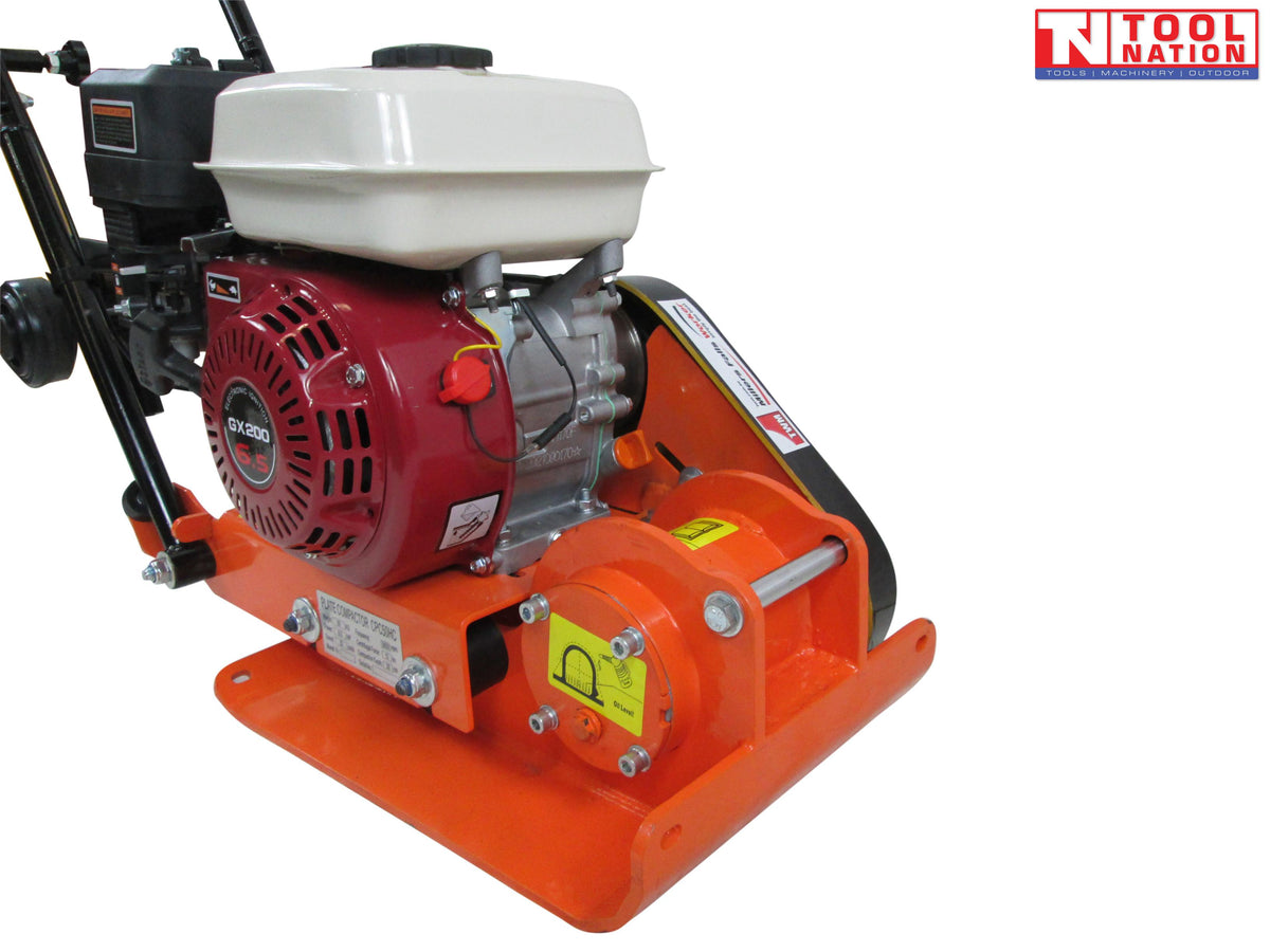 Close-up of an orange plate compactor showing the petrol engine, fuel tank, black belt cover, base plate, and warning labels.