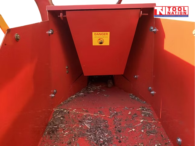 Inside view of a red feed hopper with wood chips on the floor, visible bolts, and a warning label on the centre panel.