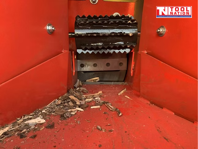 Close-up inside a red feed hopper showing black feed rollers, bolts, and wood chips on the floor.