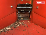 Close-up inside a red feed hopper showing black feed rollers, bolts, and wood chips on the floor.