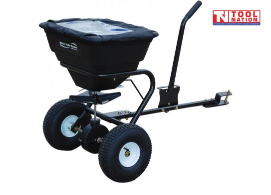 Black tow-behind spreader with a covered hopper, long drawbar, upright handle, two treaded tyres and Tool Nation logo.
