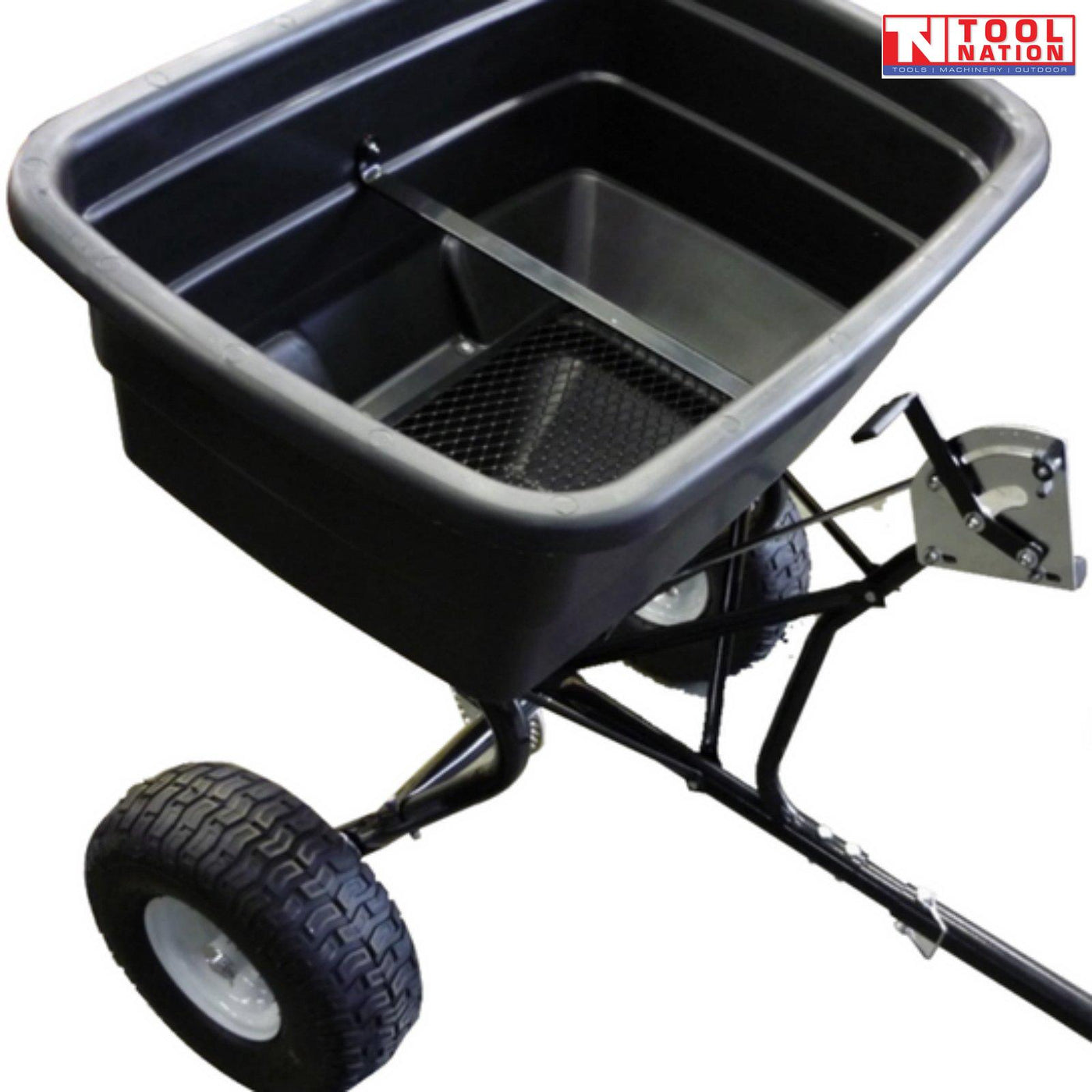 Close-up of a black tow-behind spreader showing the open hopper, mesh screen, frame, drawbar and two treaded tyres.