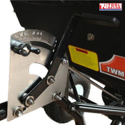 Close-up of a black tow-behind spreader showing a metal adjustment plate, black handle, bolts, warning label and part of a tyre.