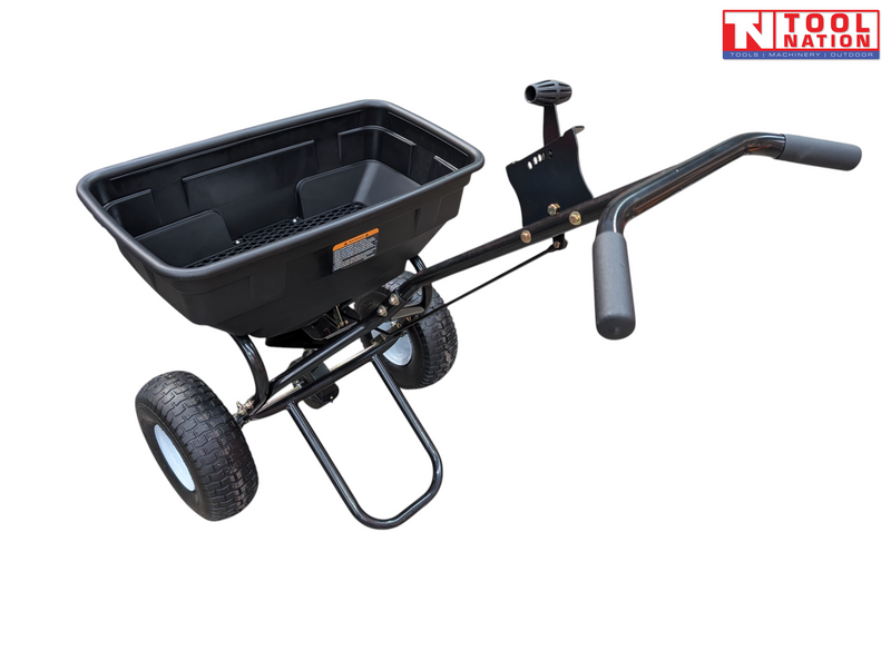 Black walk-behind spreader shown from above with an open hopper, long handle, two treaded tyres and Tool Nation logo.