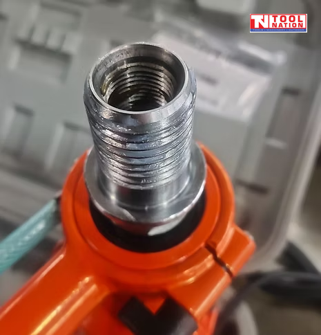Close-up of a threaded metal drill connector on an orange core drill with a hose partly visible.