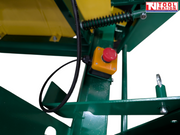 Close-up of a green and yellow saw bench frame showing a red emergency stop button, spring, cable and metal pin.