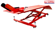 Angled front-side view of a red motorcycle lift with the rear loading ramp down, front wheel chock raised, and scissor frame partially lifted.