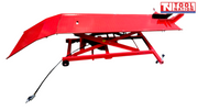 Side view of the raised red motorcycle lift with the loading ramp down, long platform, center service panel, front wheel chock, and air hose.