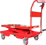 Angled view of a red hydraulic lift table cart with lowered platform, handle, wheels, foot pedal, and warning labels.